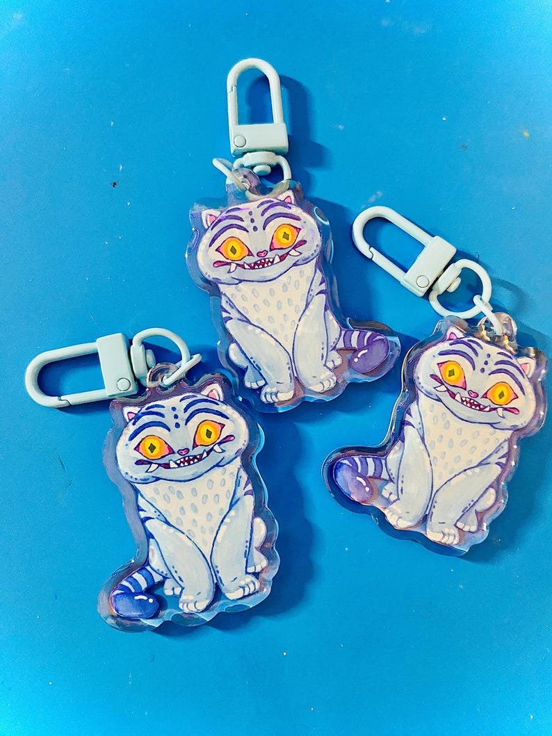 May include: Three cat-shaped keychains with light blue and white coloring, featuring yellow eyes and a purple tail. Each keychain has a light blue clasp. The keychains are on a bright blue background.