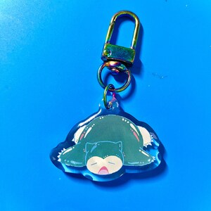 Puede incluir: Un llavero con un personaje de dibujos animados verde y azul con un cierre met&aacute;lico iridiscente arco&iacute;ris. El personaje es un Pok&eacute;mon durmiendo, Snorlax.