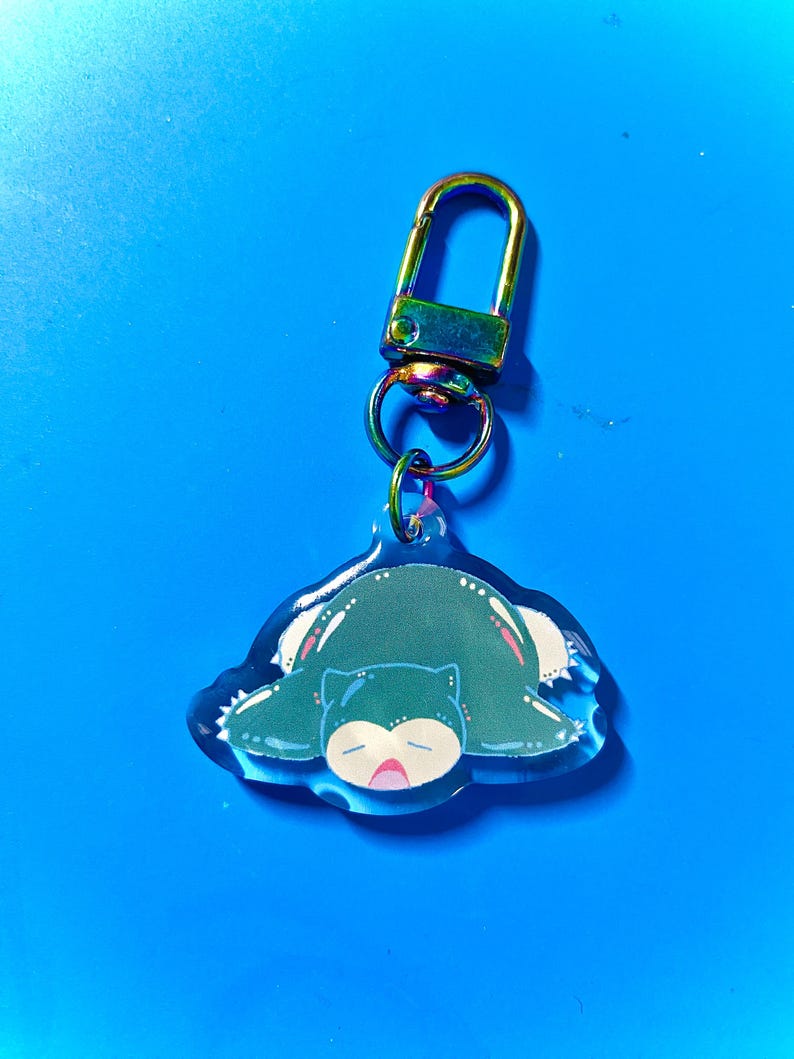 Puede incluir: Un llavero con un personaje de dibujos animados verde y azul con un cierre met&aacute;lico iridiscente arco&iacute;ris. El personaje es un Pok&eacute;mon durmiendo, Snorlax.