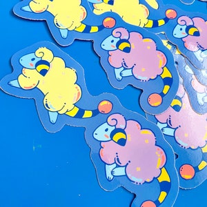 Könnte beinhalten: Ein Set bunter Schafsticker mit einem gelb-schwarz gestreiften Schwanz. Die Schafe sind in verschiedenen Posen und haben einen verspielten Gesichtsausdruck. Die Sticker sind auf blauem Hintergrund.