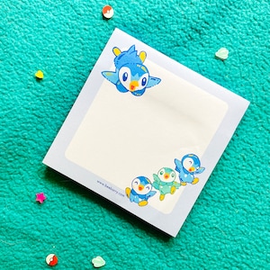 May include: A blue and white notepad with a cartoon penguin design. The notepad has a white background with blue borders and a website address printed on the bottom. The penguin design features four penguins in different poses, with one penguin flying in the air.