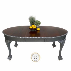 May include: A grey oval-shaped dining table with a wood top and carved legs. The table has a leaf that can be added to extend the length. The table is decorated with a rope-like design around the edge. The table is sitting on a white background.
