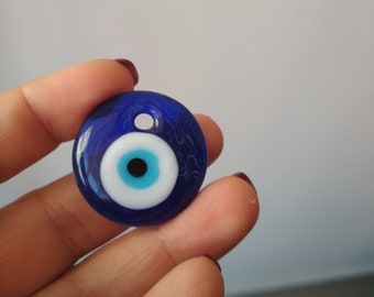 May include: A blue glass evil eye charm with a white center and a blue pupil. The charm is round and has a small hole at the top for hanging.