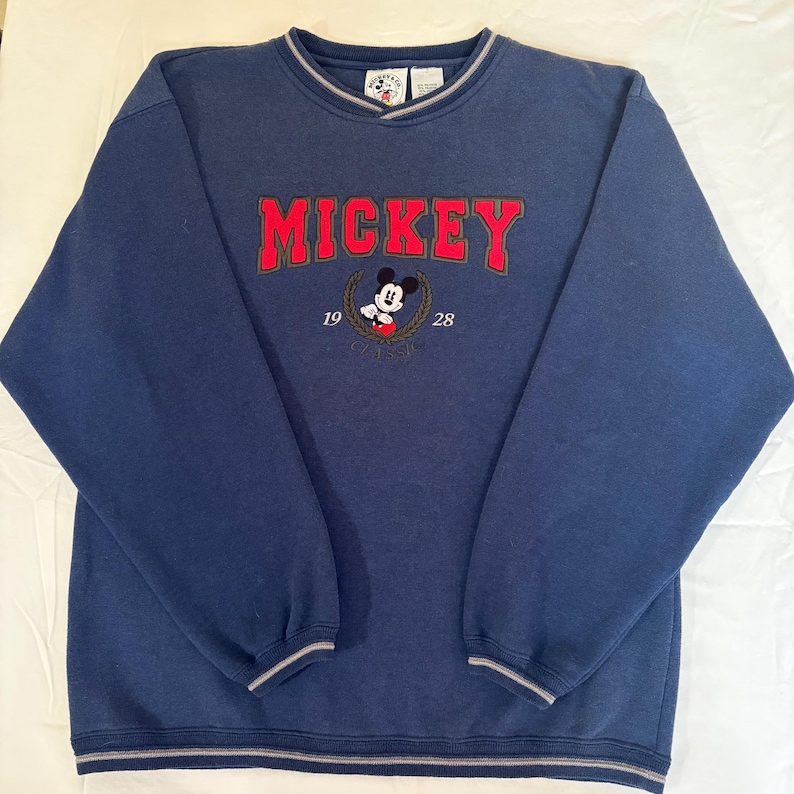 May include: A navy blue long-sleeve sweatshirt featuring the red embroidered text MICKEY above an illustration of Mickey Mouse. The design includes the numbers 19 and 28 on either side of a laurel wreath, with grey and white striped trim at the collar.