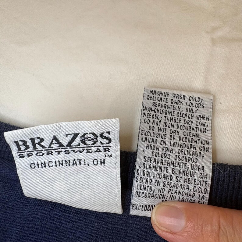 May include: A white clothing tag with the black text BRAZOS SPORTSWEAR CINCINNATI, OH and a second white care instruction tag with black text in English and Spanish, both attached to a dark navy blue fabric garment with a ribbed texture.