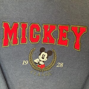 May include: A dark blue sweatshirt featuring the word MICKEY in large red embroidered letters with olive green outlines. Below, an embroidered Mickey Mouse character is centered within an olive green laurel wreath with the numbers 19 and 28.