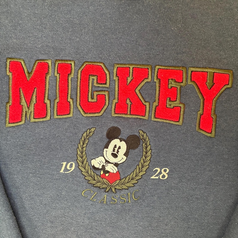 May include: A dark blue sweatshirt featuring the word MICKEY in large red embroidered letters with olive green outlines. Below, an embroidered Mickey Mouse character is centered within an olive green laurel wreath with the numbers 19 and 28.
