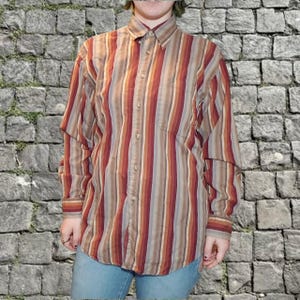May include: A button-down shirt with vertical stripes in shades of brown, red, and tan. The long-sleeved shirt has a collared neckline and a single chest pocket. The shirt is worn with blue jeans.