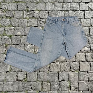 Puede incluir: Vaqueros de mezclilla azul claro con un diseño clásico de cinco bolsillos. Los vaqueros tienen un corte de pierna recta y un cierre de botón y cremallera. La mezclilla tiene una apariencia descolorida, lo que sugiere un aspecto vintage. Los vaqueros están planos sobre una superficie de piedra.