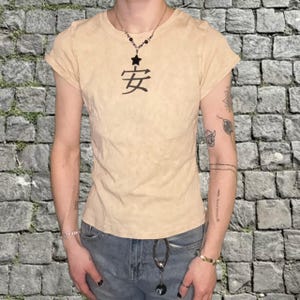 Puede incluir: Camiseta beige de manga corta con el carácter chino "安" impreso en negro. La camiseta tiene un aspecto ligeramente desgastado. El modelo lleva un collar. La camiseta es de estilo entallado.