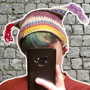May include: A colorful, handmade crochet hat with a long, pointed tip. The hat features stripes of yellow, blue, purple, and brown. A red tassel hangs from the side. The hat is a unique, whimsical accessory.