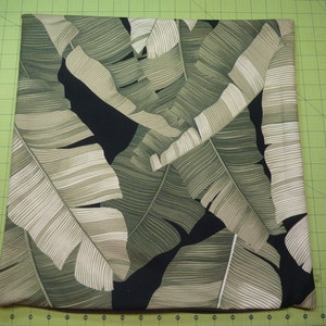 May include: A square pillow cover featuring a botanical print of large, overlapping banana leaves in shades of sage green, olive, and cream against a solid black background. The fabric has a visible texture and a repeating tropical leaf pattern.