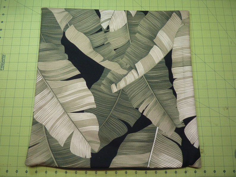 May include: A square pillow cover featuring a botanical print of large, overlapping banana leaves in shades of sage green, olive, and cream against a solid black background. The fabric has a visible texture and a repeating tropical leaf pattern.