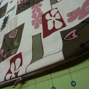 May include: A rectangular throw pillow cover featuring a cream-colored background with a repeating pattern of abstract shapes in olive green, dark brown, and muted pink. The design includes stylized floral motifs and thin lines, with a beige zipper.