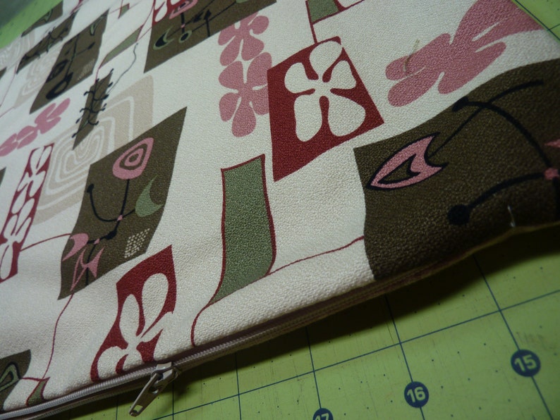 May include: A rectangular throw pillow cover featuring a cream-colored background with a repeating pattern of abstract shapes in olive green, dark brown, and muted pink. The design includes stylized floral motifs and thin lines, with a beige zipper.