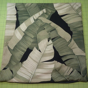 May include: A square throw pillow cover featuring a botanical print of large, overlapping banana leaves in shades of sage green, olive, and cream against a solid black background. The fabric has a visible weave and clean, straight stitched edges.