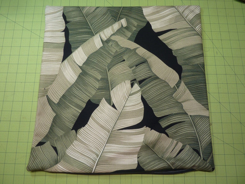May include: A square throw pillow cover featuring a botanical print of large, overlapping banana leaves in shades of sage green, olive, and cream against a solid black background. The fabric has a visible weave and clean, straight stitched edges.