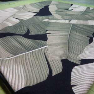 May include: A square cushion cover featuring a repeating tropical banana leaf pattern in shades of sage green, olive, and cream against a solid black background. The fabric has a visible woven texture and includes a black zipper along the side edge.
