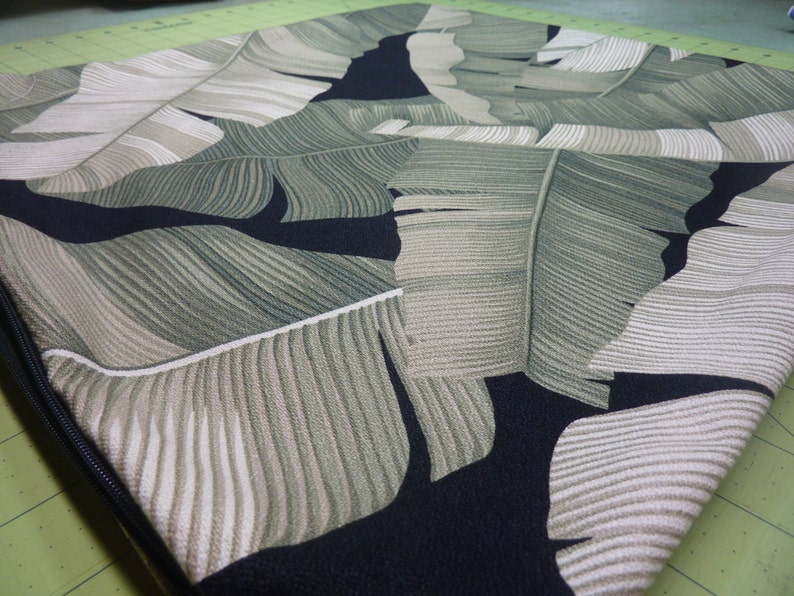 May include: A square cushion cover featuring a repeating tropical banana leaf pattern in shades of sage green, olive, and cream against a solid black background. The fabric has a visible woven texture and includes a black zipper along the side edge.