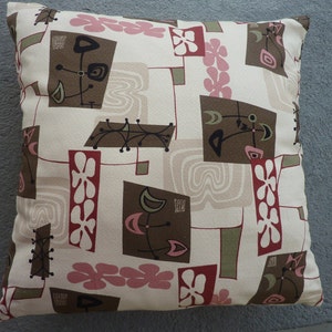 May include: A square throw pillow featuring a cream-colored background with a repeating pattern of brown rectangles, pink flower shapes, and olive green accents. The design includes black line art motifs and light beige wavy line patterns.