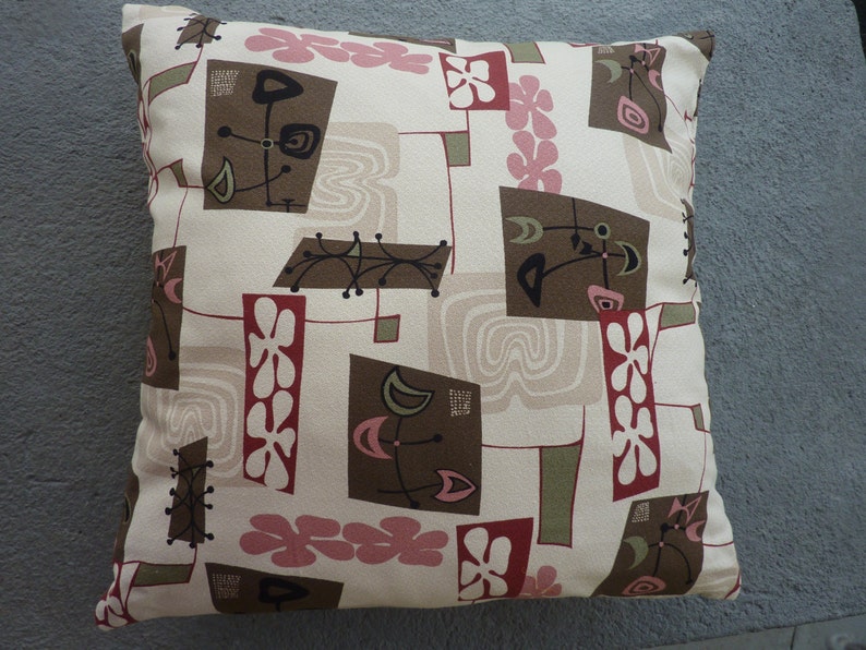 May include: A square throw pillow featuring a cream-colored background with a repeating pattern of brown rectangles, pink flower shapes, and olive green accents. The design includes black line art motifs and light beige wavy line patterns.