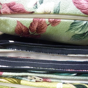 May include: A stack of fabric cushion covers featuring various floral patterns in shades of green, red, and white. Each cover includes a visible zipper closure, with some zippers in a light beige tone and others in a contrasting black color.