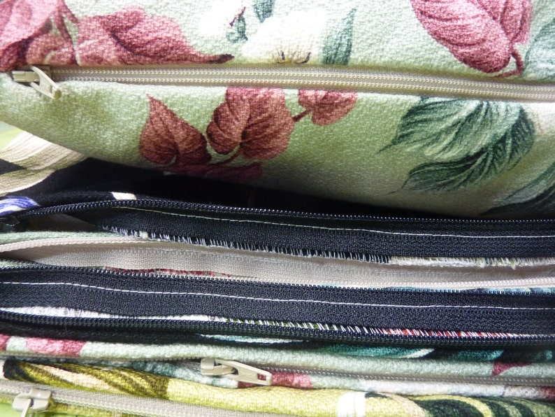 May include: A stack of fabric cushion covers featuring various floral patterns in shades of green, red, and white. Each cover includes a visible zipper closure, with some zippers in a light beige tone and others in a contrasting black color.
