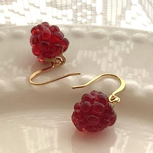 May include: A pair of red raspberry-shaped earrings with gold-colored ear wires. The earrings are made of a translucent red material, and the raspberry shapes have a textured surface. The earrings are displayed on a white, textured surface.