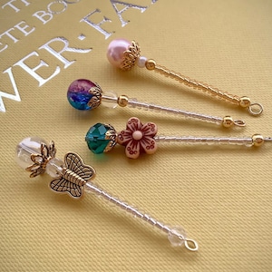 May include: Four decorative bookmarks with different charms. The charms include a butterfly, a flower, a green bead, and a pink pearl. The bookmarks are made with clear and gold beads.