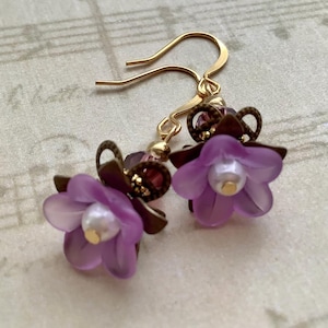 May include: A pair of delicate floral earrings. Each earring features a translucent purple flower with a pearl-like center, accented by dark brown filigree and gold-colored ear wires. The earrings are set against a background of sheet music.