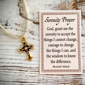 May include: A gold cross necklace with a white ribbon. The necklace comes with a card that reads "Serenity Prayer" and includes the text "God, grant me the serenity to accept the things I cannot change, courage to change the things I can, and the wisdom to know the difference. -Reinhold Niebuhr".