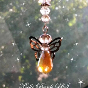 May include: A beaded angel car charm with a bronze-colored metal angel silhouette and a large, faceted amber-colored crystal pendant. The charm is suspended from a chain of small, dark beads. The text "Belle Beads US" is at the bottom.