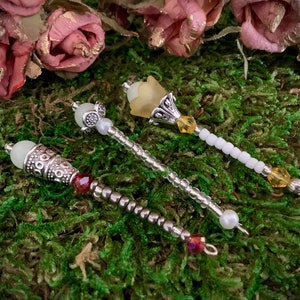 May include: Three decorative wands with different colored beads and charms. The wands are made of silver metal and have a variety of beads, including white, red, and clear. The charms are a silver flower, a silver bell, and a silver star.