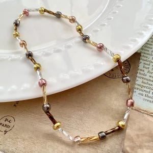 May include: A delicate necklace featuring a mix of metallic beads in gold, silver, bronze, and pink. The beads are strung on a twisted wire, creating a unique and elegant design. The necklace is displayed on a white plate.
