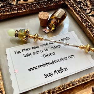 Könnte beinhalten: Ein kleiner, silberner und goldener Zauberstab mit einer grün-goldenen Perle an einem Ende. Der Zauberstab liegt auf einer weißen Karte mit dem Text "Tip: Place mini wand under a bright light source to "charge" it. -Theresa www.bellebeadsus.etsy.com Stay Magical".