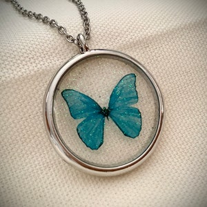 May include: A silver chain necklace with a round pendant featuring a blue butterfly encased in clear resin with glitter.