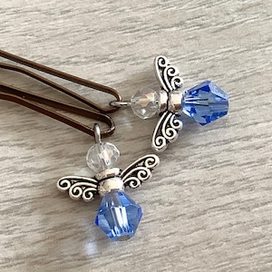 May include: Two silver angel charms with blue crystal accents. The charms are attached to bobby pins.