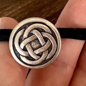 Op de afbeelding: Een zilverkleurige haarband met een Keltisch knoopontwerp. Het ronde, decoratieve element is bevestigd aan een zwarte elastische band. Het knoopontwerp is gedetailleerd en verhoogd, waardoor het een driedimensionale uitstraling heeft. De haarband is een stijlvol accessoire.