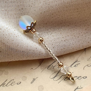 May include: A handcrafted, gold-toned wand-shaped charm with a pearlescent orb at the top. The charm features clear and gold beads, with a delicate filigree design. The wand is designed for jewelry making or as a decorative element.