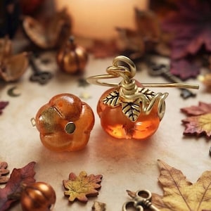 May include: Two orange pumpkin-shaped charms, one featuring a metallic gold finish and the other topped with a gold-toned metal stem and leaf detail. The charms are crafted from a translucent material and display a smooth, rounded, segmented surface.