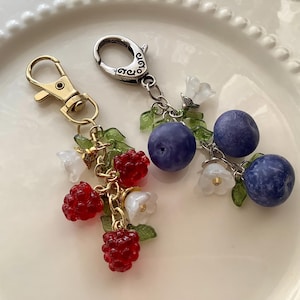 Op de afbeelding: Twee sleutelhangers met fruit- en bloemhangers. Eén sleutelhanger heeft rode frambozenhangers, witte bloemenhangers en groene bladerenhangers. De andere sleutelhanger heeft blauwe bosbessenhangers, witte bloemenhangers en groene bladerenhangers. Beide hebben metalen sluitingen.