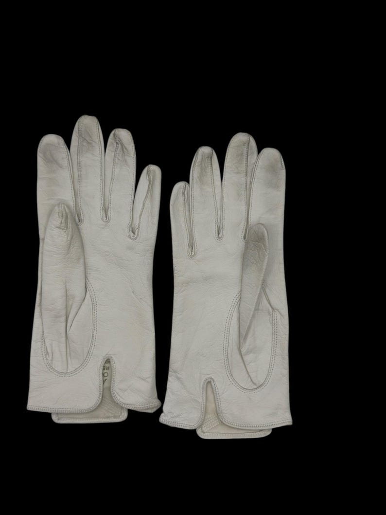 May include: A pair of off-white leather gloves. The gloves have a classic design with individual finger slots and a split at the wrist. The stitching is visible, and the gloves appear to be well-worn.