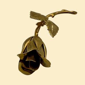 May include: A gold-colored metal rose sculpture with detailed petals and leaves. The rose is in full bloom, with a curved stem and two leaves extending from the side. The sculpture has a vintage aesthetic.