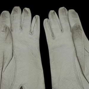 May include: A pair of off-white work gloves, likely leather, with visible stitching and a textured surface. The gloves are set against a black background, highlighting their shape and detail. The gloves appear to be new.