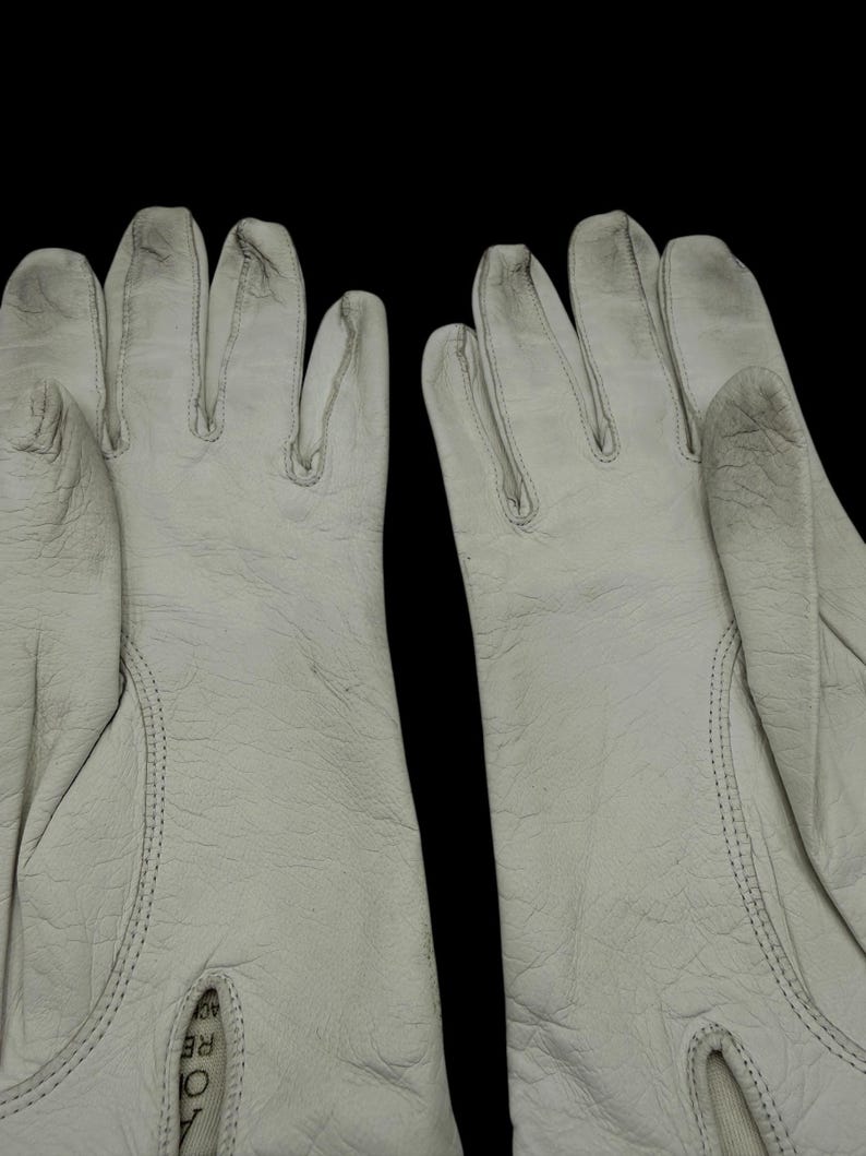 May include: A pair of off-white work gloves, likely leather, with visible stitching and a textured surface. The gloves are set against a black background, highlighting their shape and detail. The gloves appear to be new.