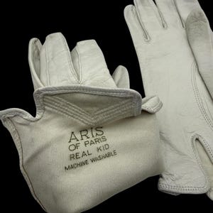 May include: A pair of off-white leather gloves with the text "ARIS OF PARIS REAL KID MACHINE WASHABLE" printed on the cuff. The gloves are set against a black background, highlighting their texture and design.