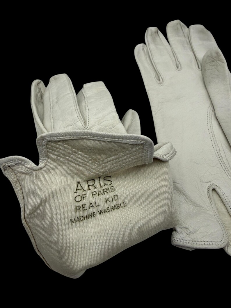 May include: A pair of off-white leather gloves with the text "ARIS OF PARIS REAL KID MACHINE WASHABLE" printed on the cuff. The gloves are set against a black background, highlighting their texture and design.