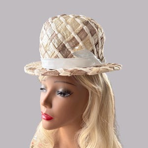 Könnte beinhalten: Ein Vintage-Hut mit einem gewebten Muster in Creme und Braun. Der Hut hat eine runde Krone und eine breite Krempe, akzentuiert mit einem weißen Band. Dieses stilvolle Accessoire ist perfekt, um einen Hauch von Retro-Flair zu verleihen.