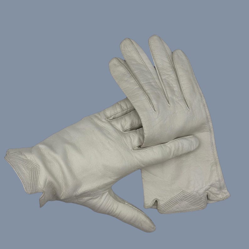 May include: A pair of off-white leather gloves. The gloves are shown with the fingers slightly curved, and the cuffs have a decorative stitched design. The gloves are made of a soft, supple leather.