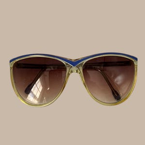 May include: Vintage sunglasses with a light brown gradient lens and a gold frame. The top of the frame is accented with a blue stripe. The sunglasses have a classic, retro style.